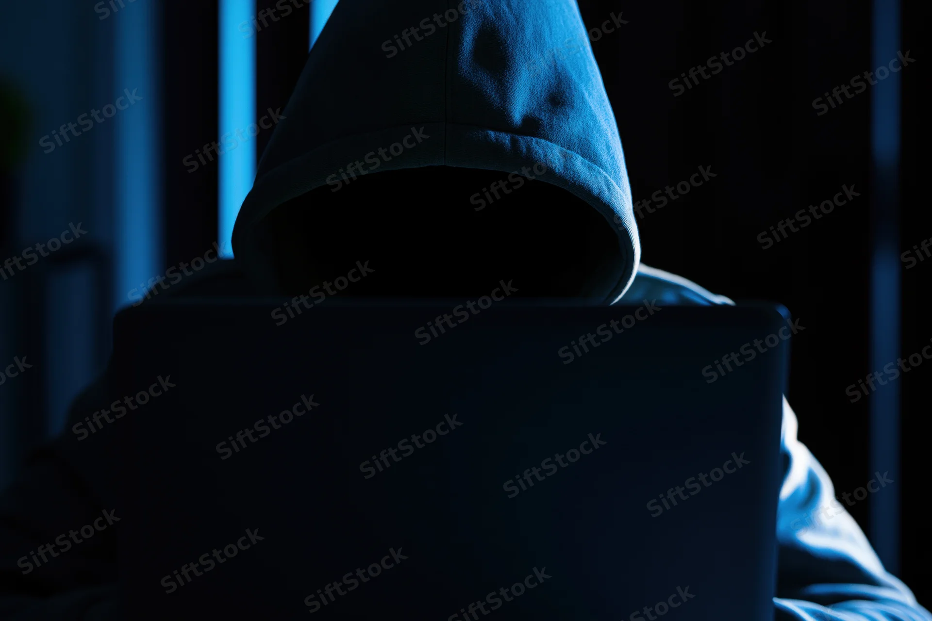 Hooded figure sitting behind laptop in dark room with blue glow