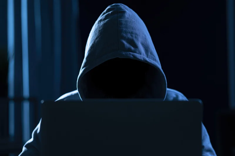 Hooded figure sitting behind laptop in dark room