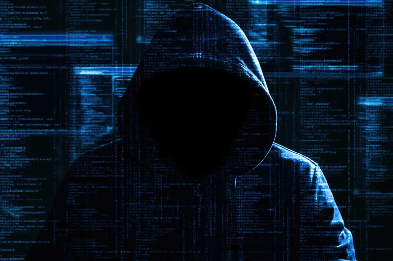 Hooded figure in shadow against glowing blue code display