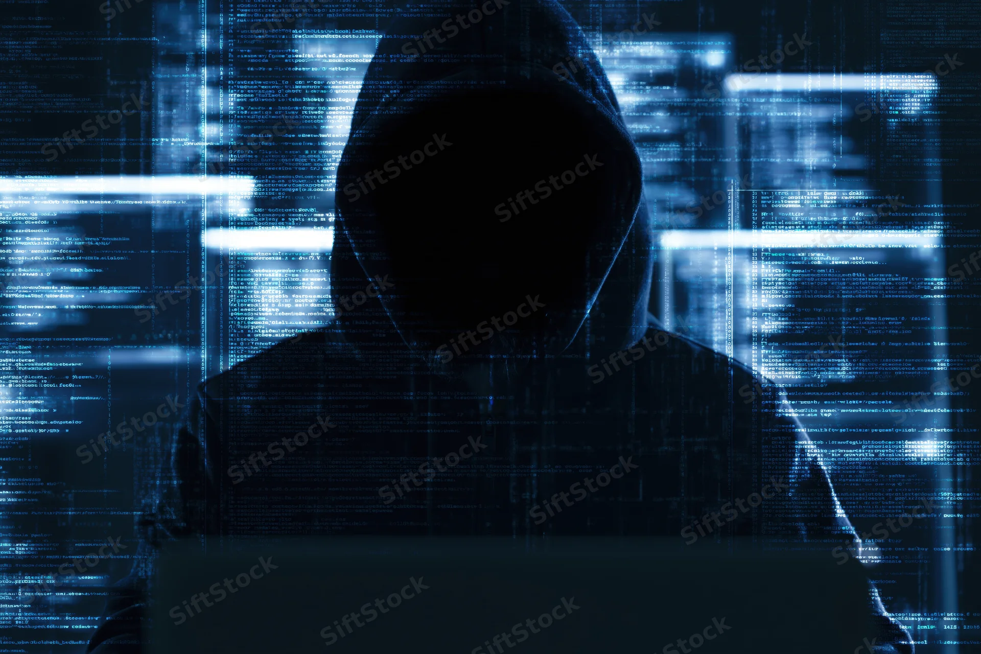 Hooded figure in silhouette using laptop with digital code overlay in dark environment