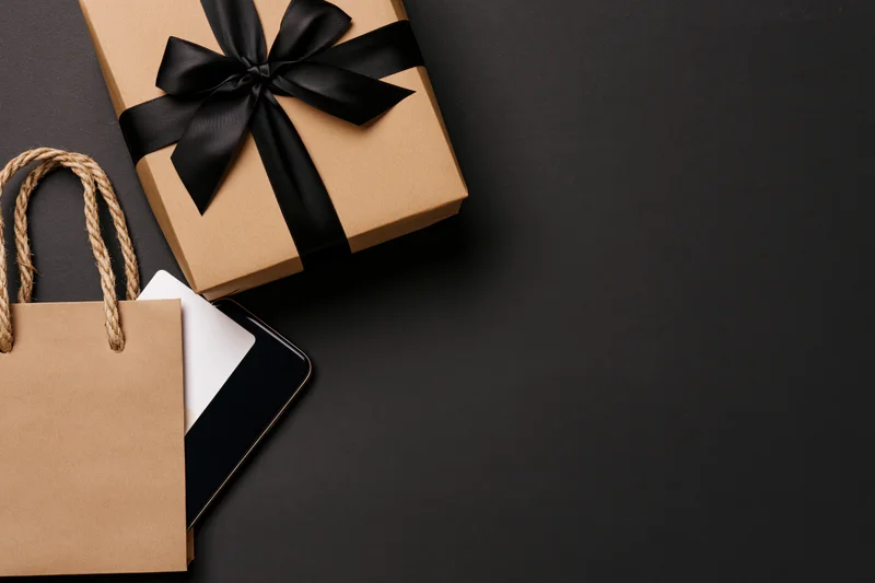 Kraft paper gift box with black satin bow and shopping bag beside smartphone on dark surface
