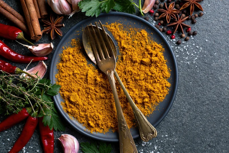 Ground turmeric powder on a dark plate with fork and spoon surrounded by whole spices and herbs