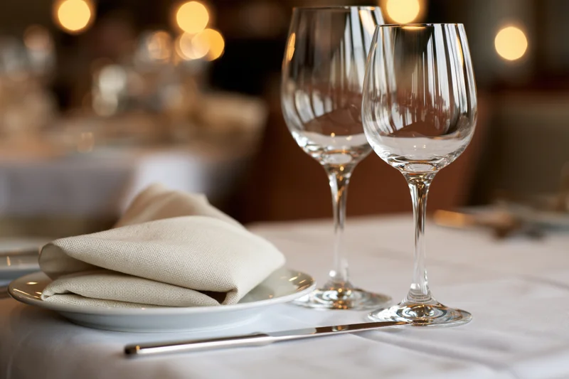 Two empty wine glasses and a folded linen napkin on a white plate at a formally set dining table in a restaurant
