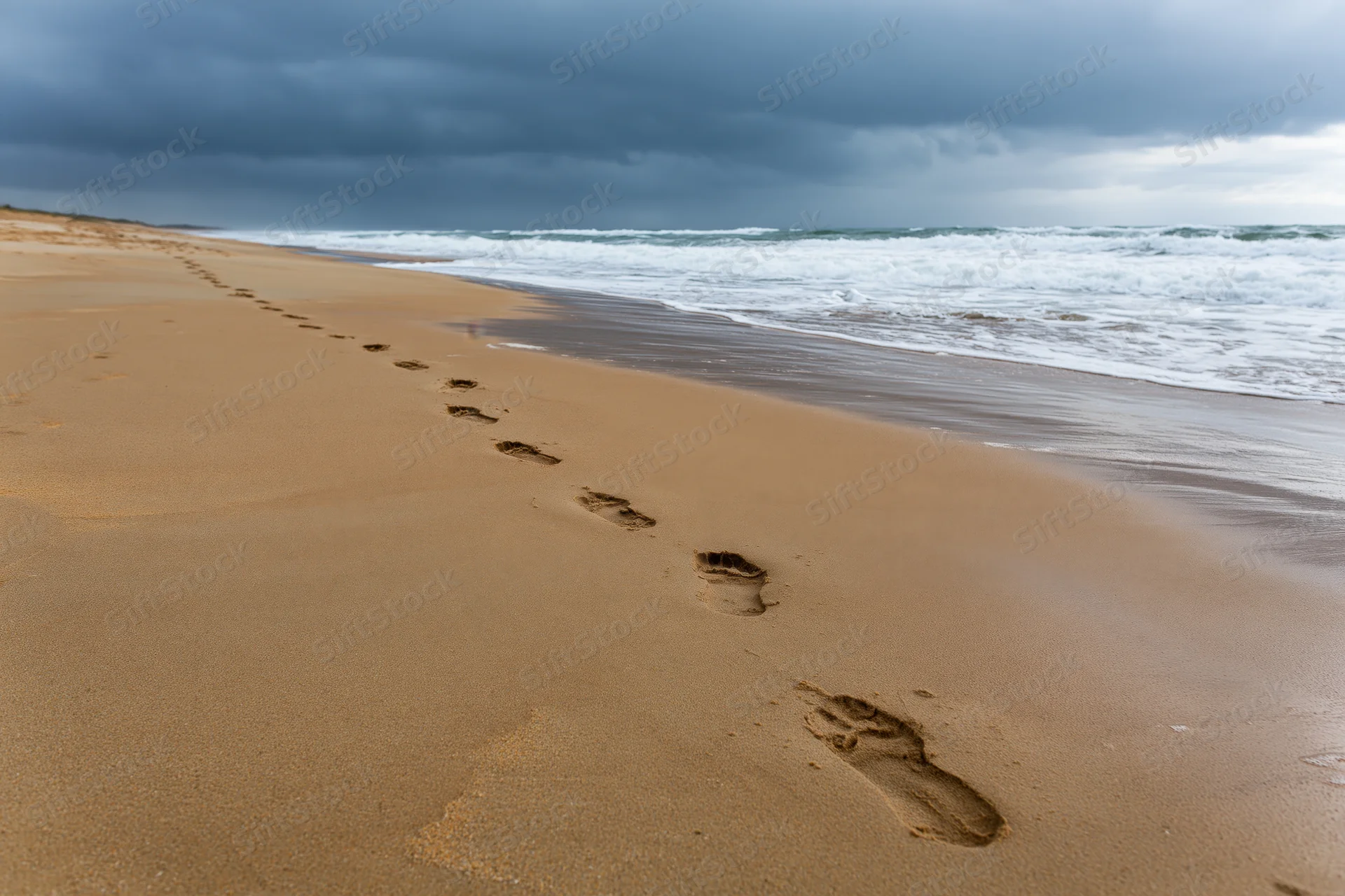 Footprints in wet amber sand leading toward the horizon along the beach under a stormy sky