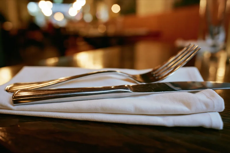 Fork and knife resting on a folded white napkin atop a dark wooden table in a restaurant setting