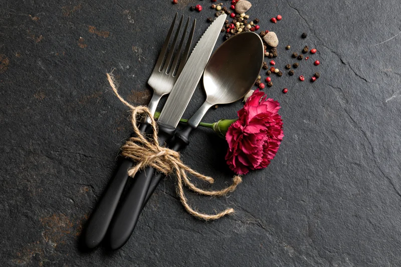 Fork knife and spoon tied with twine and a red carnation on dark slate surface with scattered peppercorns