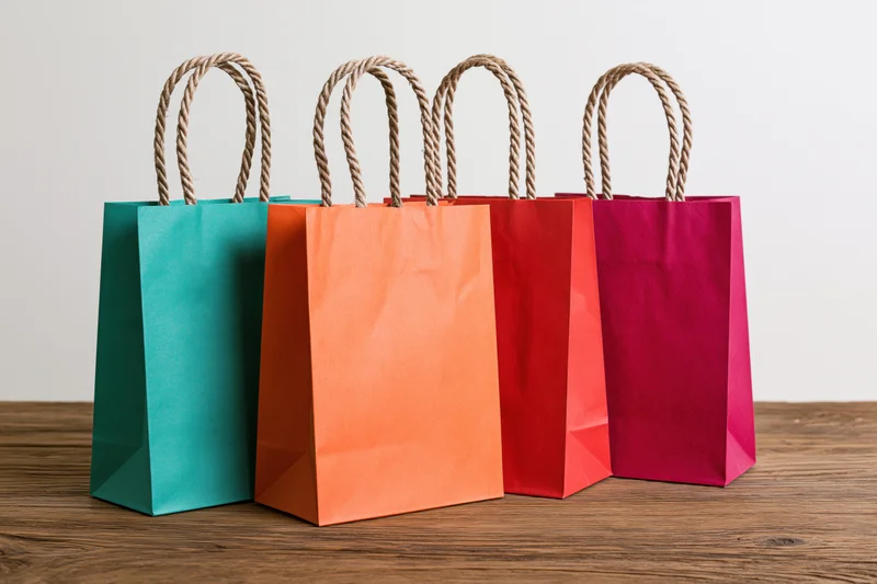 Four colorful paper shopping bags with twisted rope handles arranged in a row on a wooden table