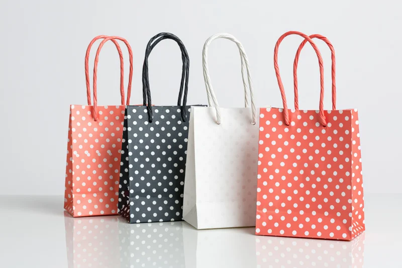 Four small paper gift bags with white polka dots arranged in a row on a reflective white surface