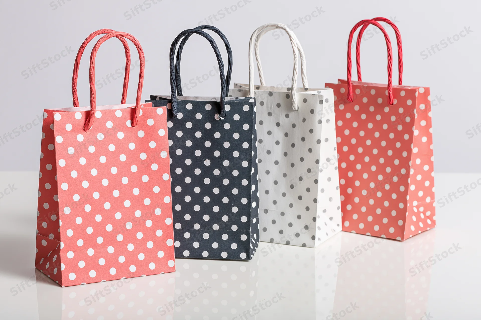 Four small paper gift bags with white polka dots arranged in a row on a reflective white surface
