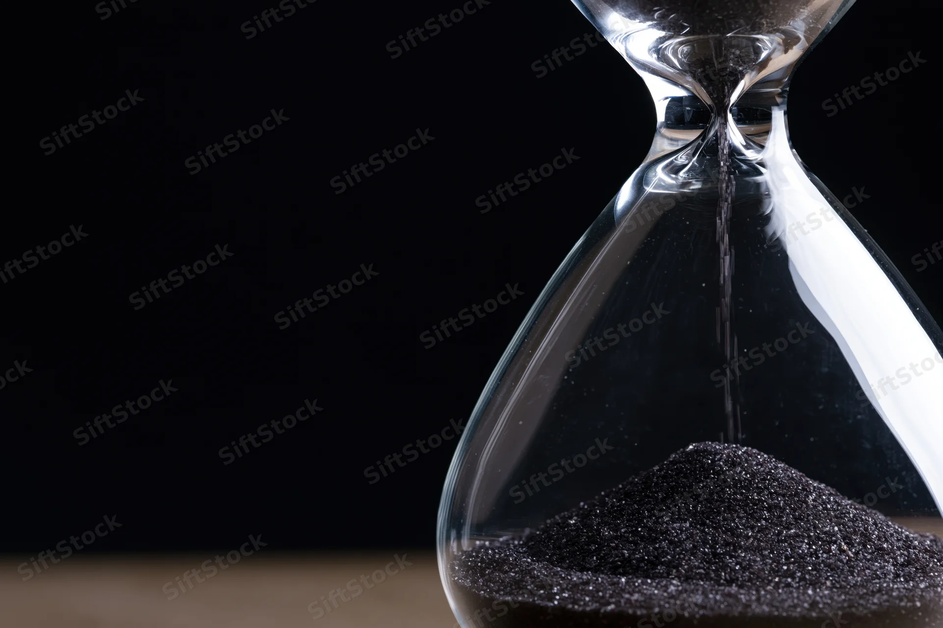 Close-up of a glass hourglass with dark sand trickling through the narrow neck against a black background