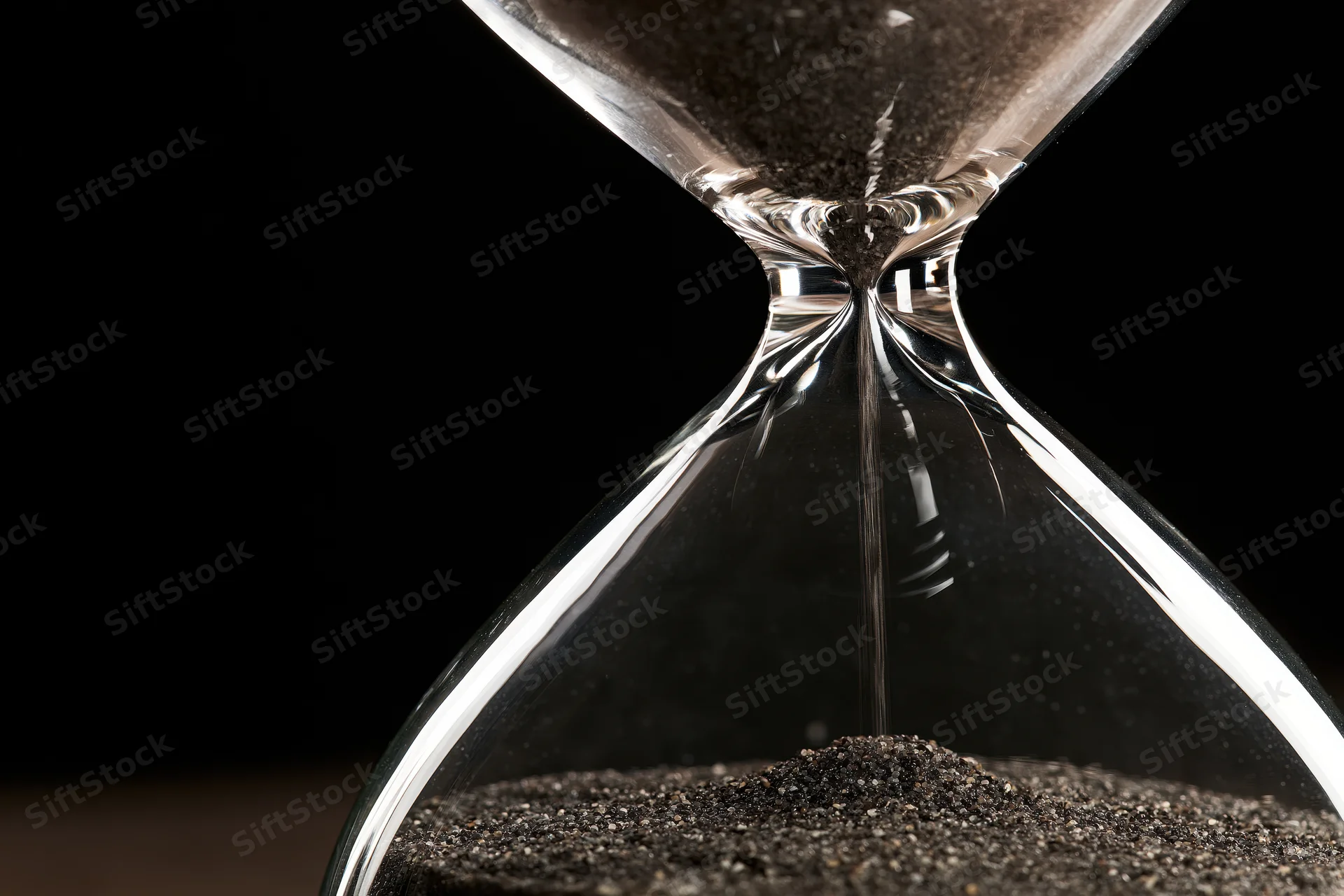 Closeup of a glass hourglass with dark sand trickling through the narrow neck against a black background