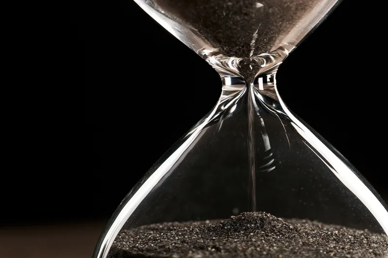 Closeup of a glass hourglass with dark sand trickling through the narrow neck against a black background
