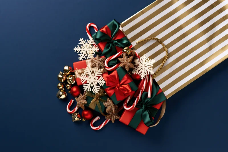 Gold and white striped shopping bag filled with wrapped gifts and christmas ornaments on blue background