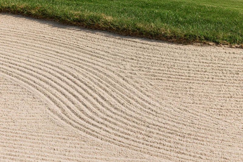 Raked sand bunker with curved parallel lines bordering green grass on a golf course