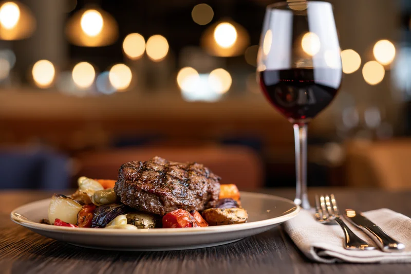 Grilled steak with roasted vegetables on a plate beside a glass of red wine on a wooden table in a dimly lit restaurant