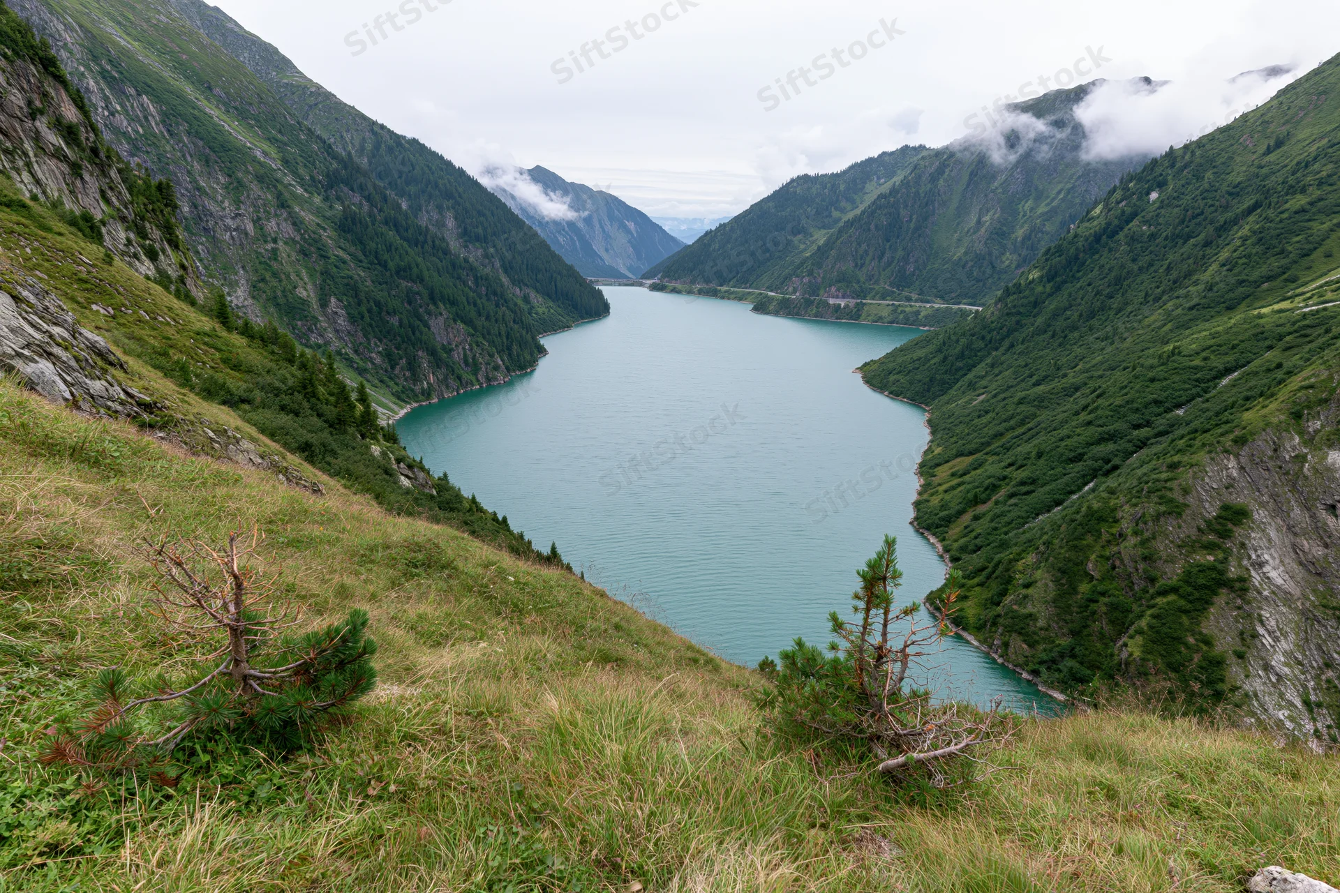 High-altitude reservoir nestled between steep green mountains, viewed from grassy slope above