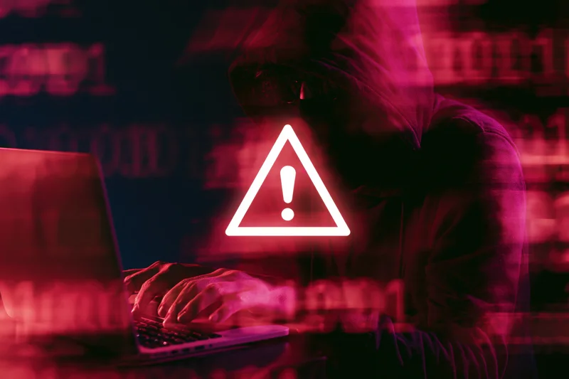 Hooded figure typing on laptop with glowing warning symbol in dark red digital environment