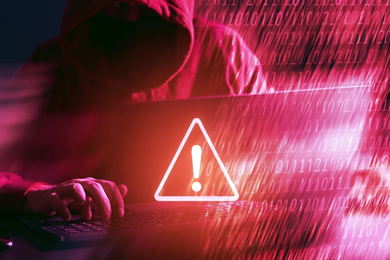 Hooded hacker typing on laptop with warning symbol and binary code stream in red light