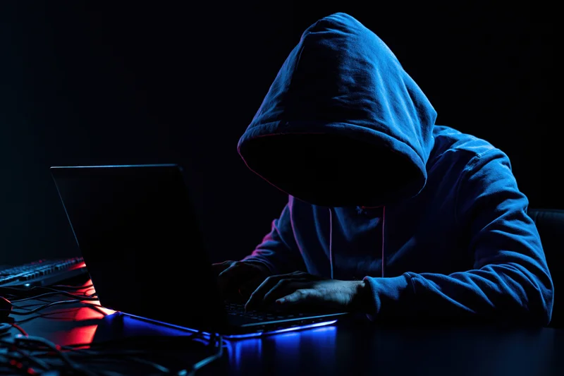 Hooded figure typing on laptop at dark desk in blue and red light