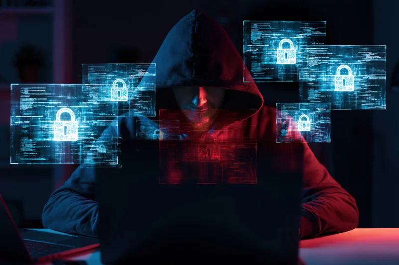 Hooded figure working on laptop with digital security holograms in dark room