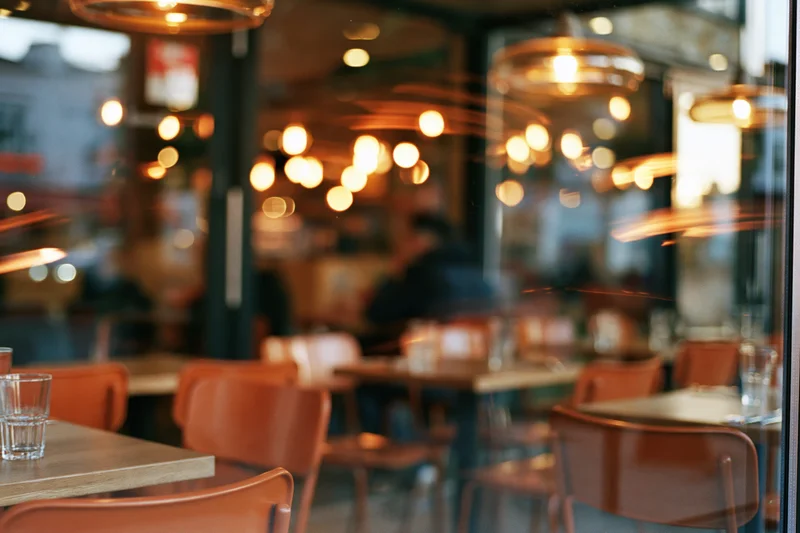 Interior of a casual restaurant viewed through glass with wooden tables and chairs, warm pendant lights glowing in the background