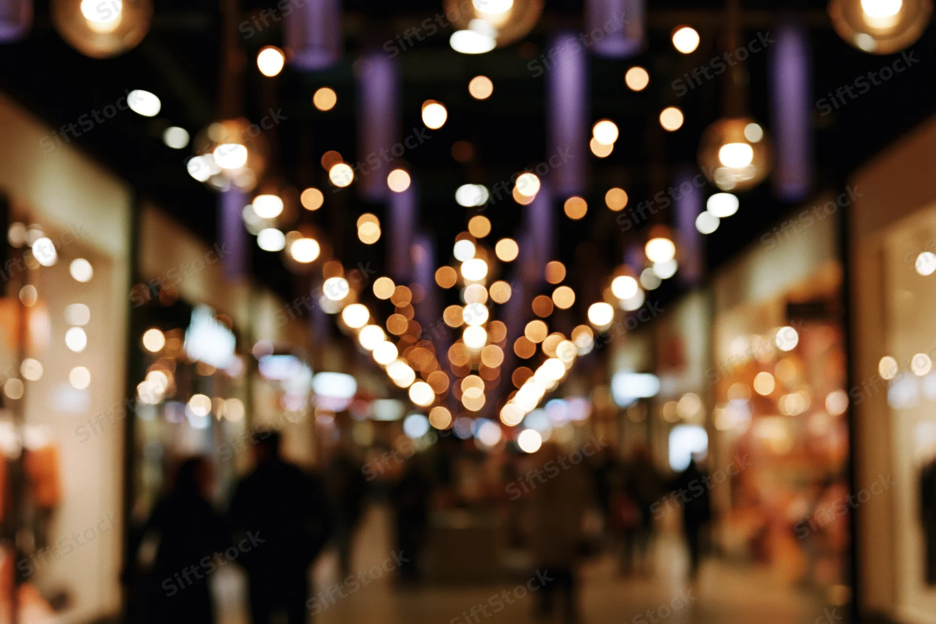 Blurred view of a shopping mall corridor at night with warm pendant lights and silhouetted people walking