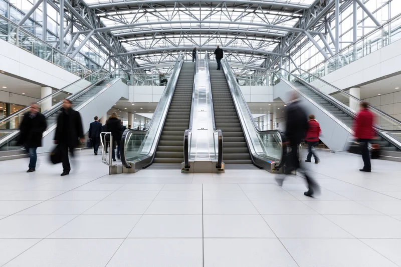 People moving through a modern airport atrium with twin escalators under a glass ceiling