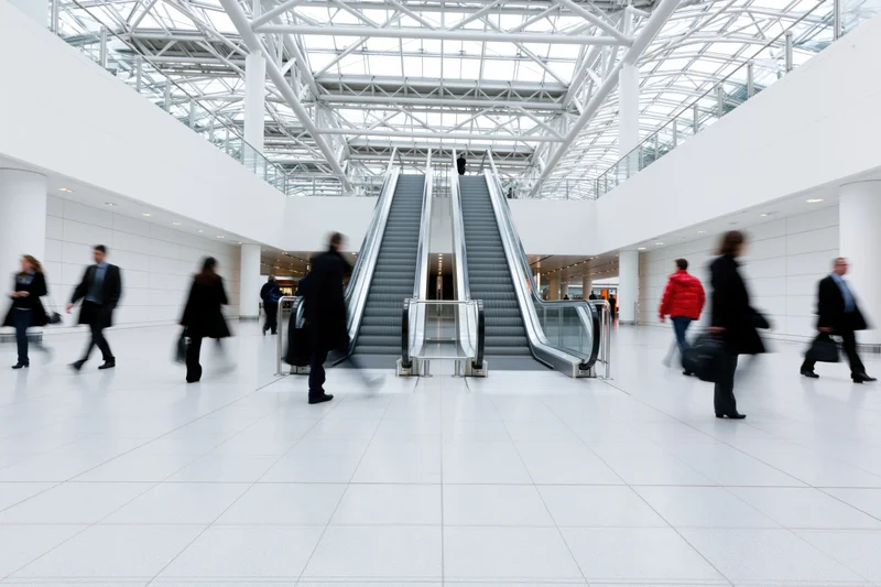 People walking through a modern airport atrium with twin escalators under a glass ceiling