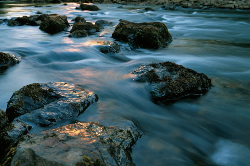 Long-exposure view of flowing river water around moss-covered rocks in shallow stream at golden hour