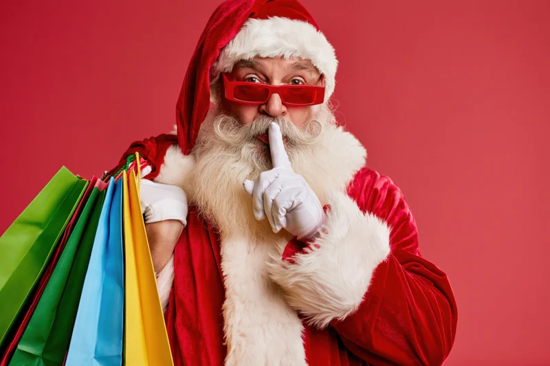 Santa in red costume holding colorful shopping bags with finger to lips against red background