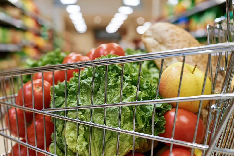 Metal shopping cart filled with fresh groceries including lettuce, tomatoes, apple and bread in a supermarket aisle