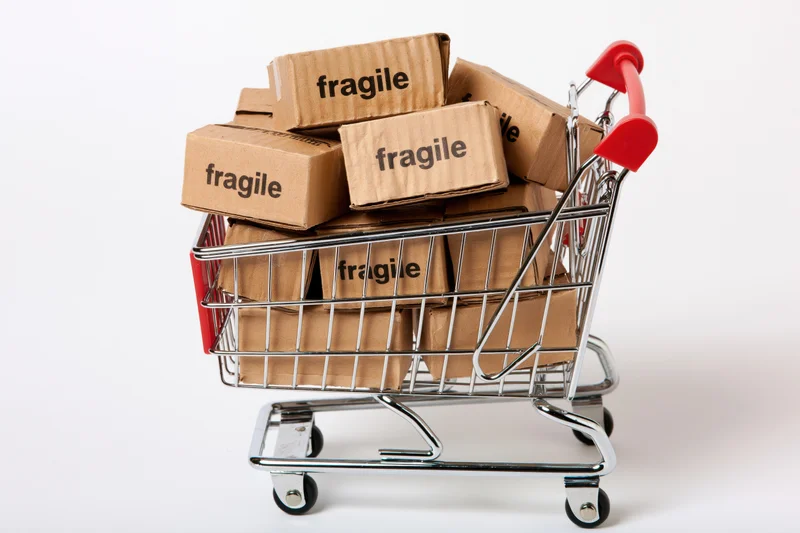 Miniature chrome shopping cart filled with small cardboard boxes labeled fragile on white background