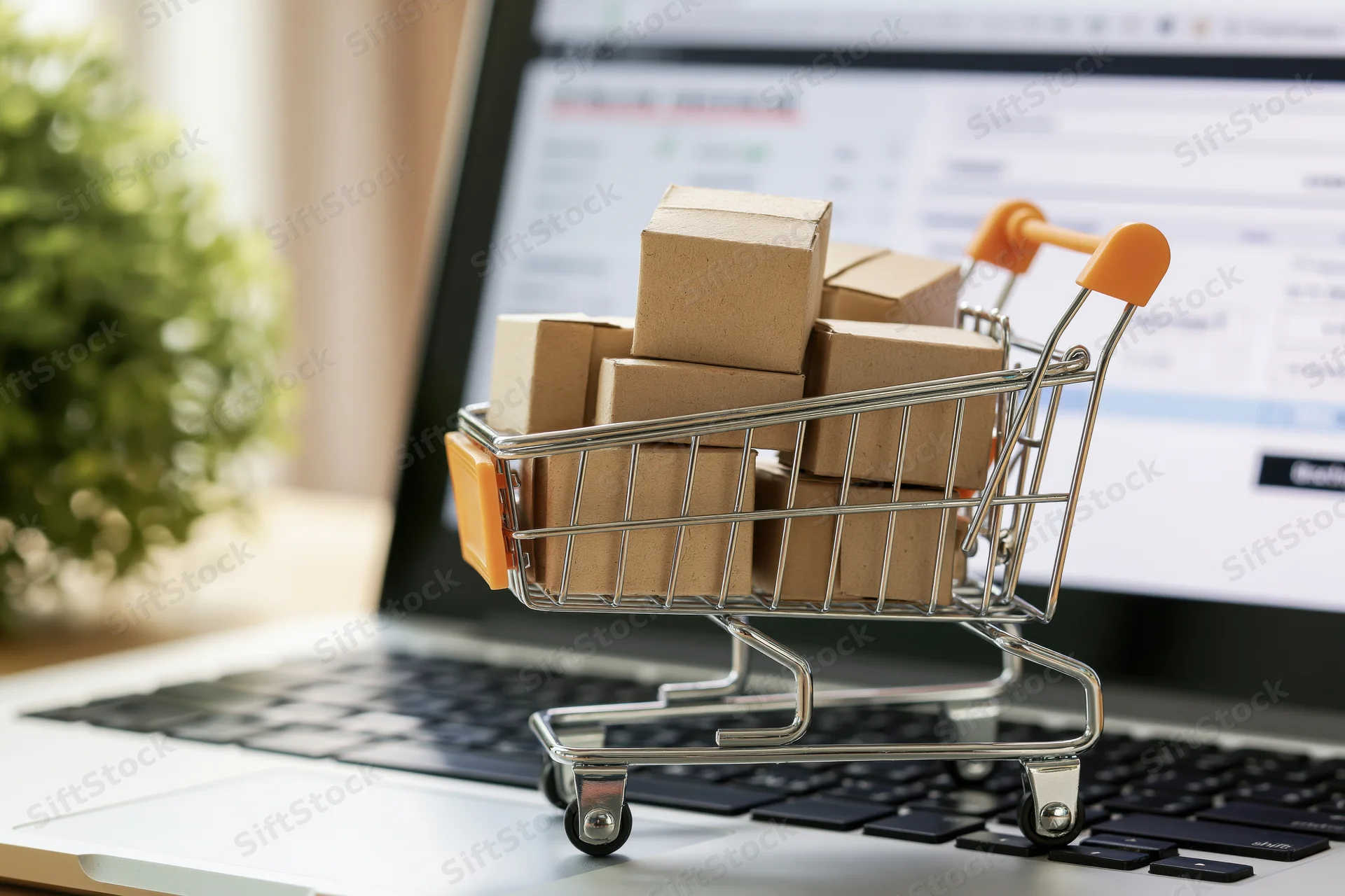 Miniature metal shopping cart filled with small cardboard boxes on laptop keyboard in front of online store screen