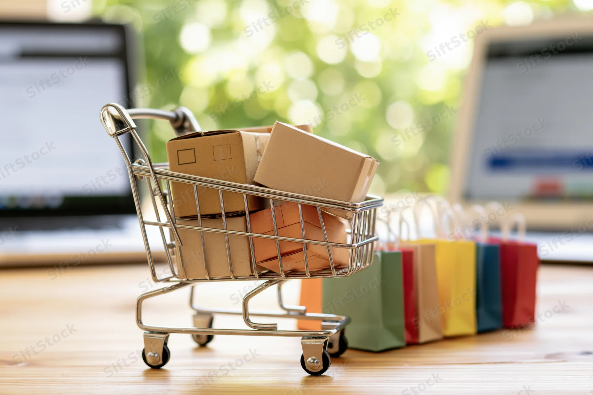 Miniature metal shopping cart filled with small cardboard boxes on wooden desk with laptops in background