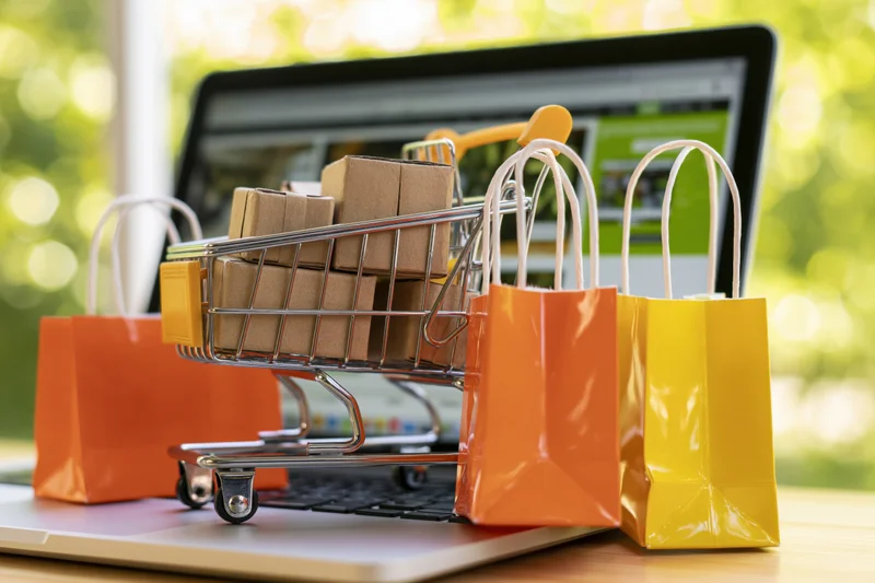 Miniature shopping cart filled with small cardboard boxes on a laptop keyboard with colorful paper bags beside it