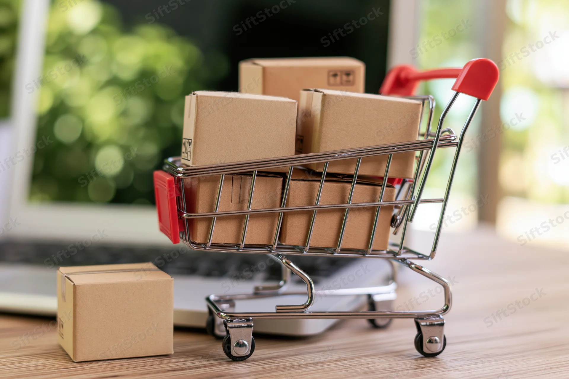 Miniature shopping cart filled with cardboard boxes on a desk beside a laptop