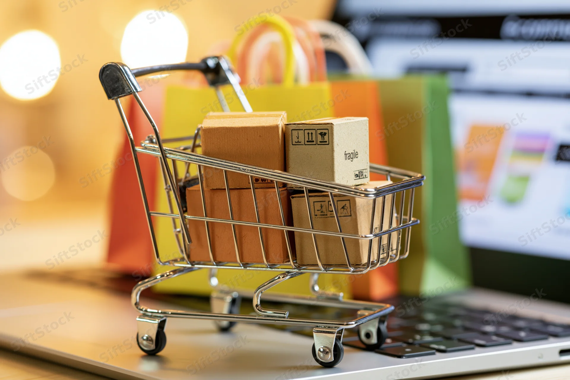 Miniature metal shopping cart filled with small cardboard boxes on a laptop keyboard with colorful bags and screen in background