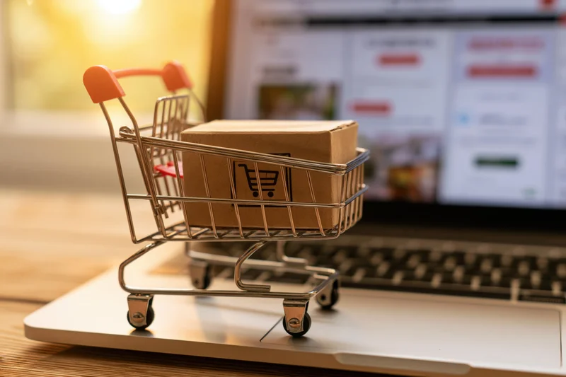Miniature metal shopping cart holding a cardboard box on a laptop keyboard with a blurred online store in the background
