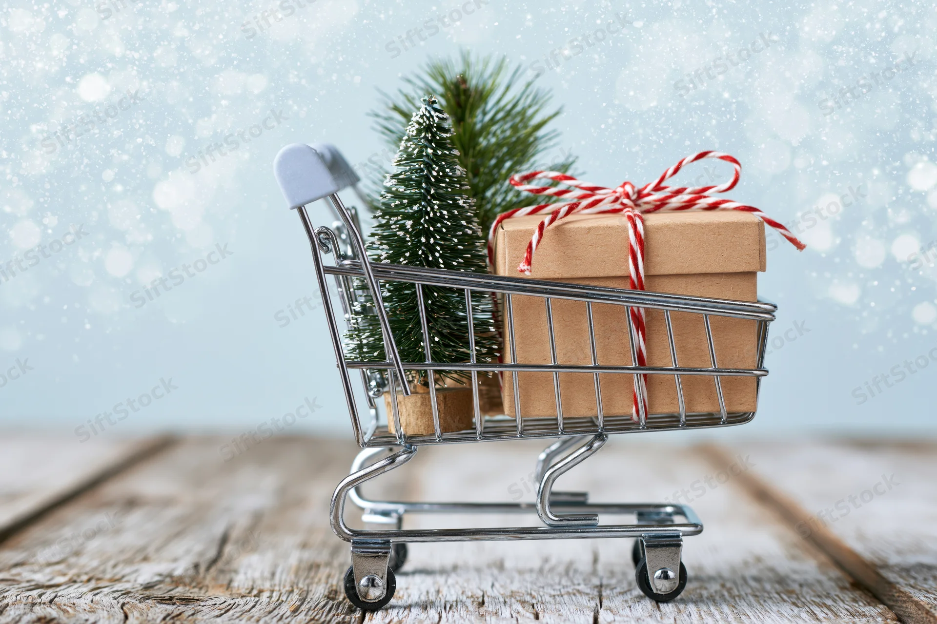 Miniature shopping cart with wrapped gift and small pine tree on wooden surface during snowy winter scene