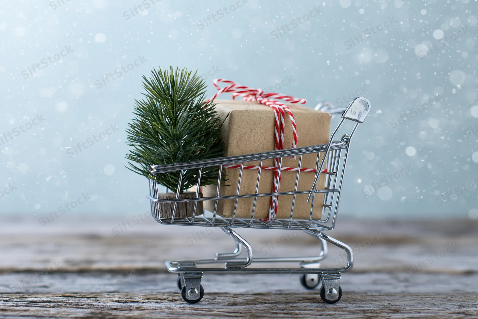 Miniature shopping cart holding a wrapped gift and pine branch on wooden surface with falling snow