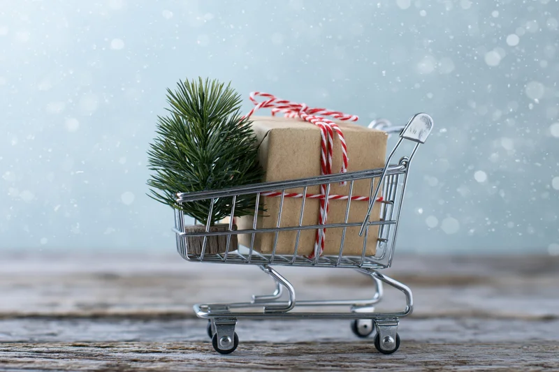 Miniature shopping cart holding a wrapped gift and pine branch on wooden surface with falling snow