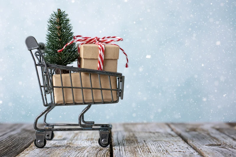 Miniature metal shopping cart holding a small decorative pine tree and wrapped gift on a wooden surface with falling snow