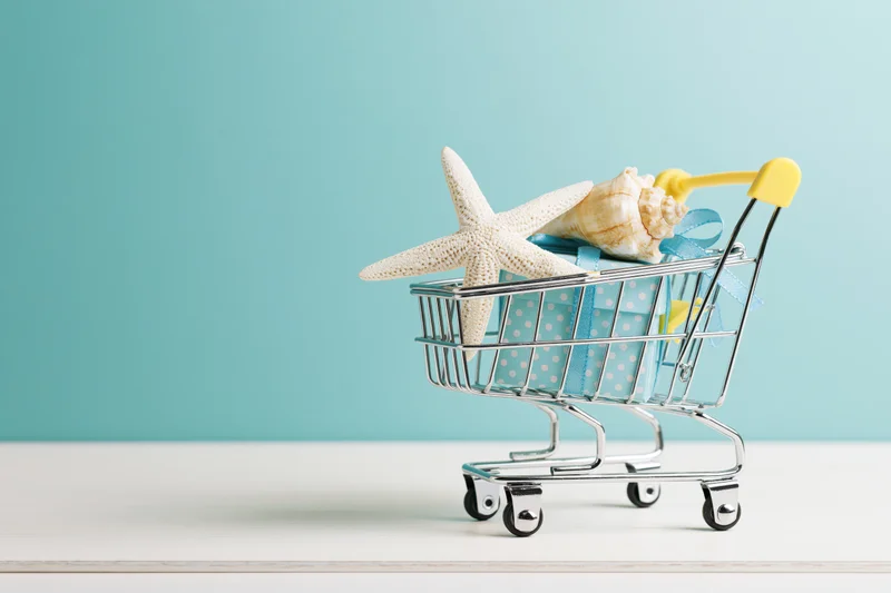 Miniature metal shopping cart holding a starfish and seashell on a gift box with ribbon, set against a turquoise background