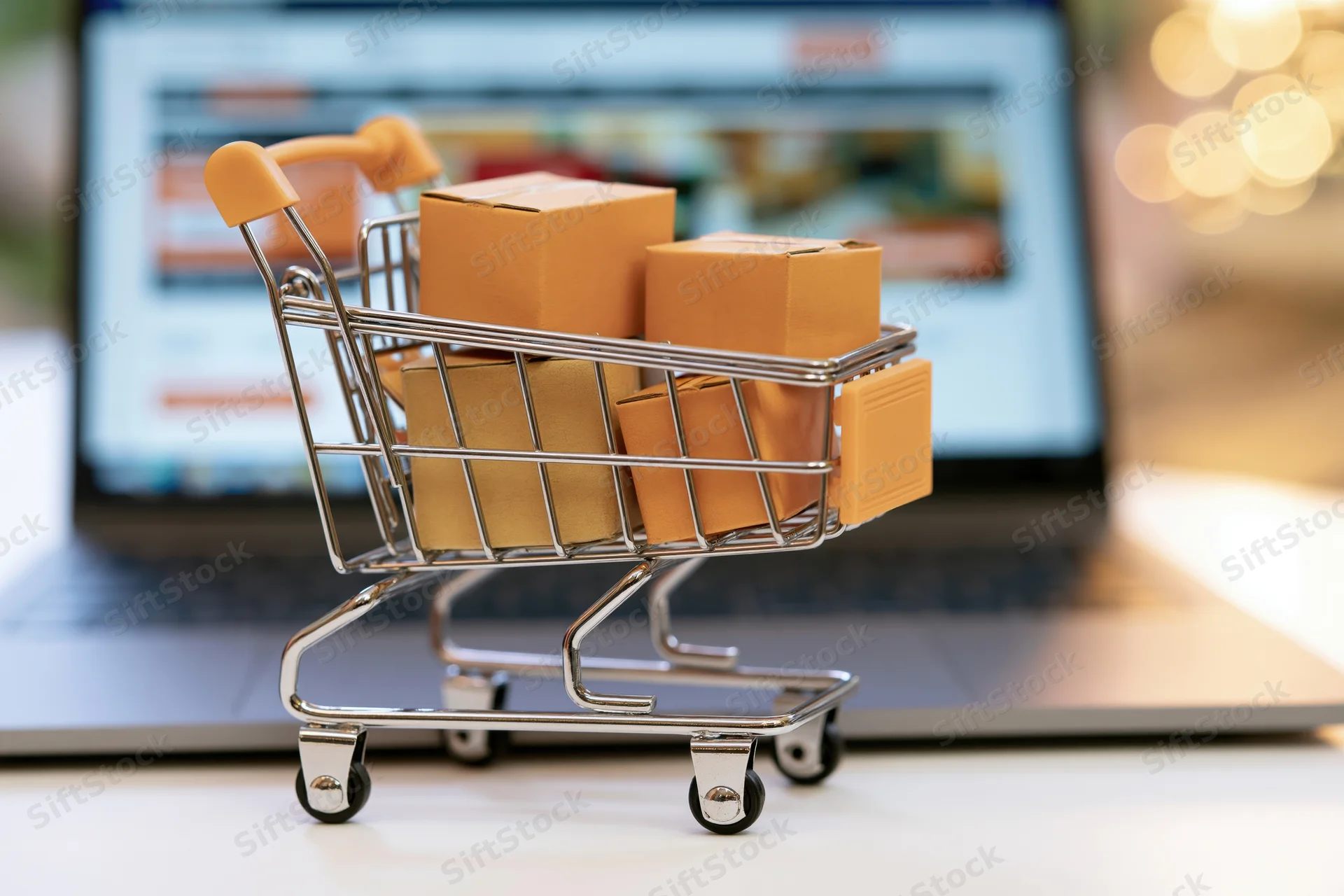 Miniature shopping cart filled with cardboard boxes in front of a laptop screen displaying an online store