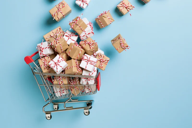 Miniature metal shopping cart overflowing with small wrapped gift boxes on blue background
