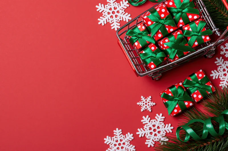 Miniature shopping cart filled with wrapped gifts and holiday decor on red background