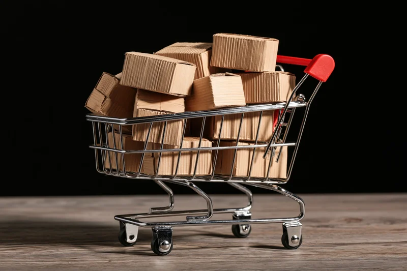 Miniature metal shopping cart overflowing with cardboard boxes on wooden surface against black background