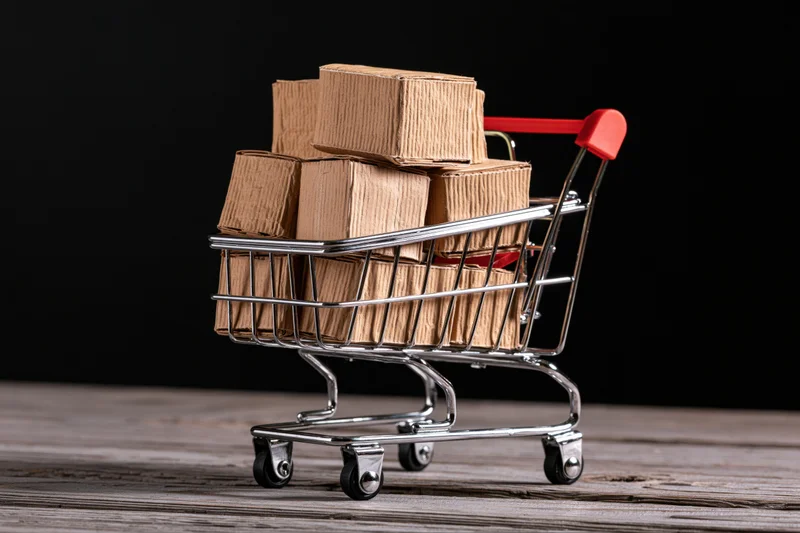 Miniature metal shopping cart overflowing with cardboard boxes on wooden surface