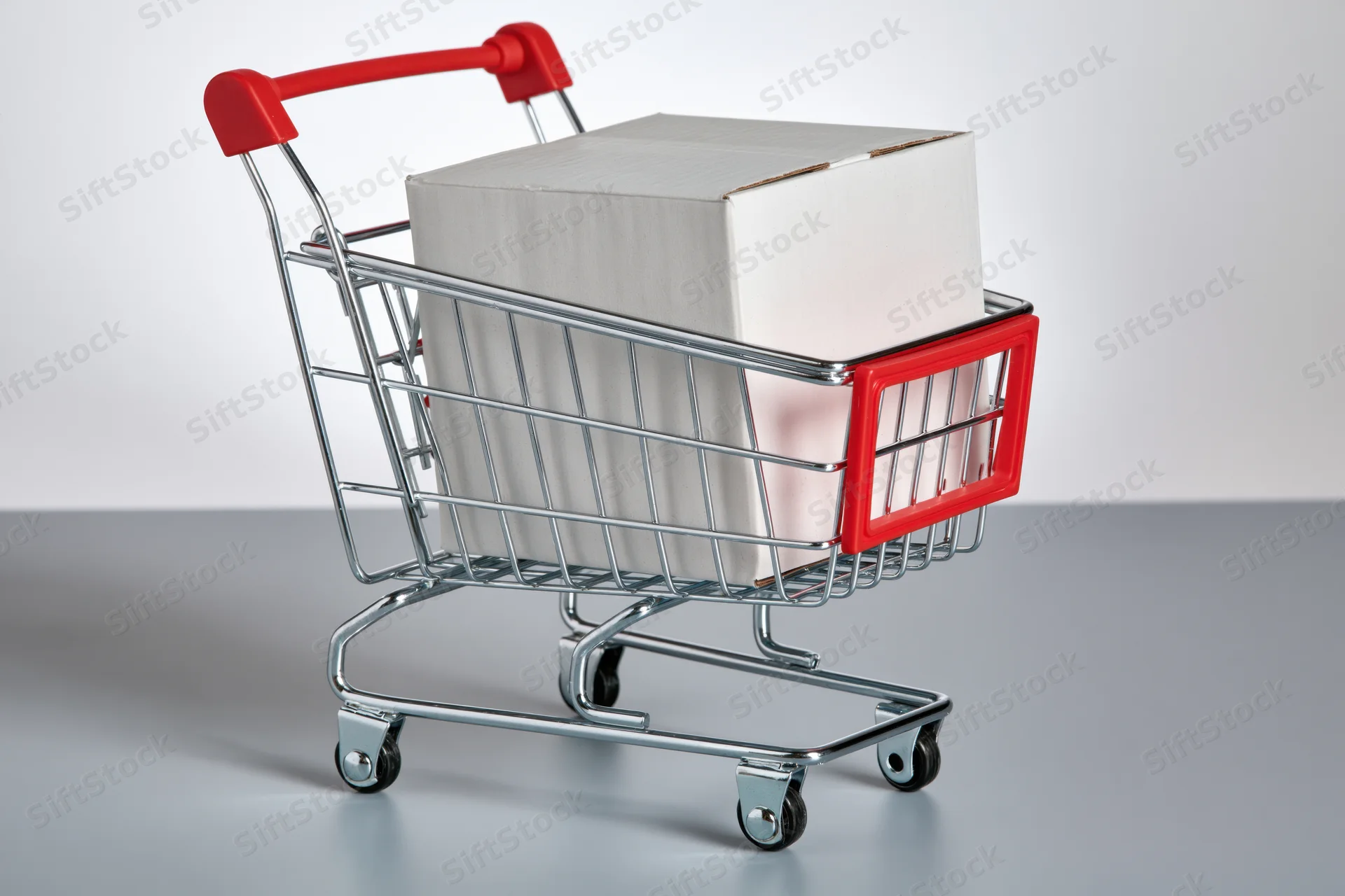 Miniature metal shopping cart with red plastic trim holding a cardboard box on a gray surface