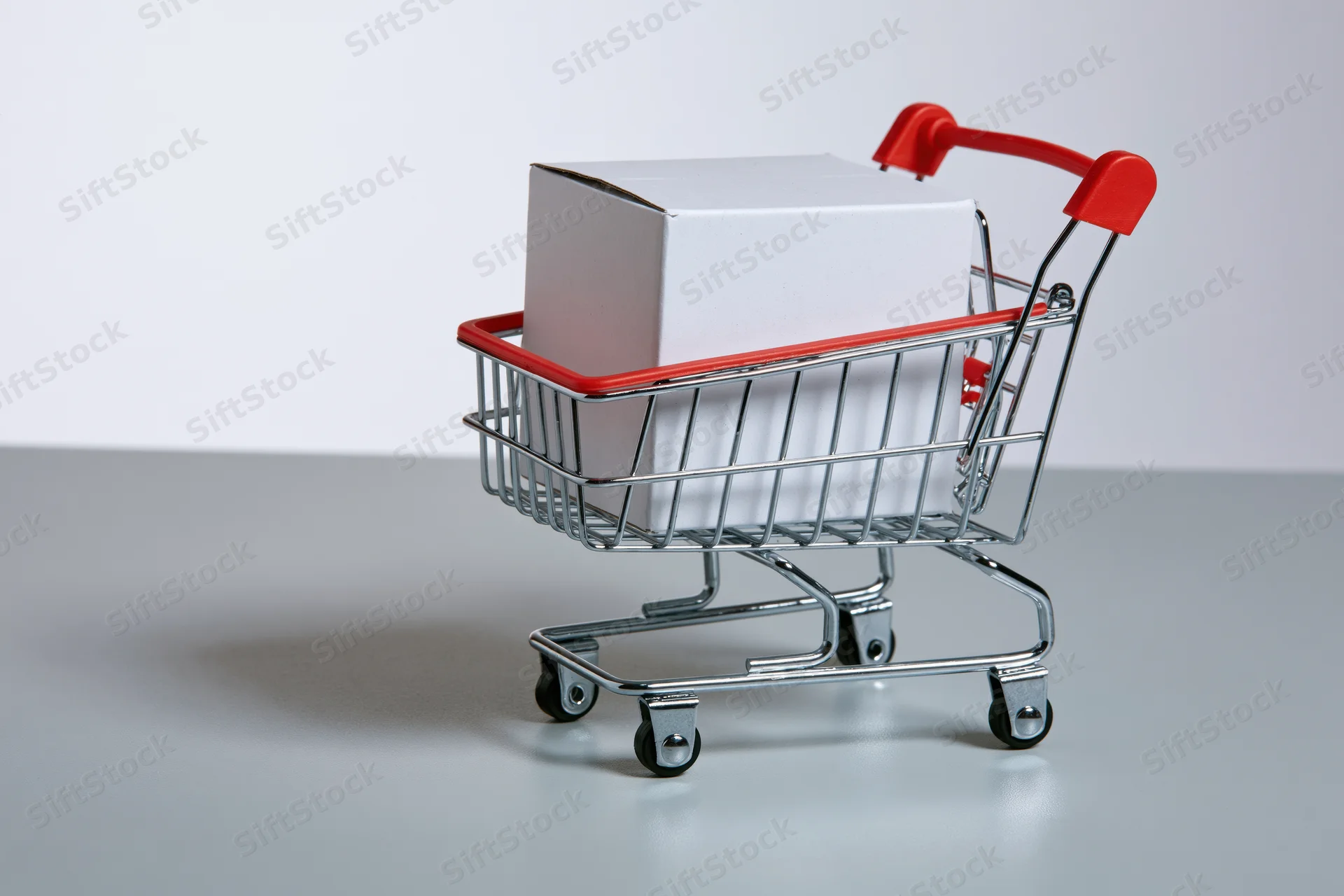 Miniature metal shopping cart with red plastic trim holding a white box on a gray surface